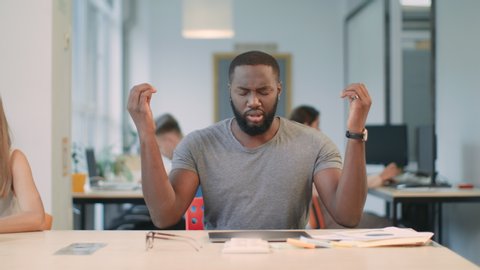Young man getting bad news at workplace in coworking space. Upset business man closing laptop computer in office. African guy meditating at workplace. Tired man doing yoga exercise in office