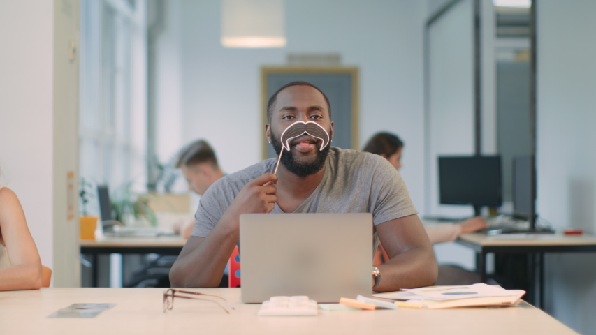 African man working on laptop computer at coworking. Joyful man looking camera and making male with moustache symbol in office. Cheerful man having fun sitting on notebook at workplace. | Shutterstock HD Video #1031923772