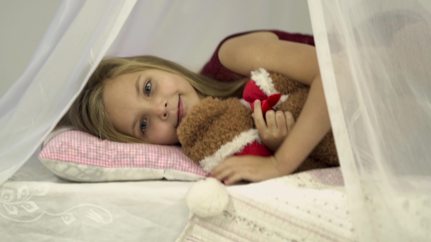 The small girl dressed in red is lying in her soft snowy cozy bed with a teddy bear that wears santa hat. Standing up the girl