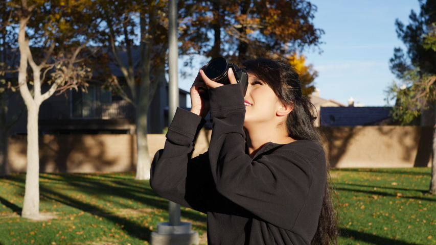 A female photographer happy and smiling in autumn leaves at sunset taking photos with a digital camera in slow motion.