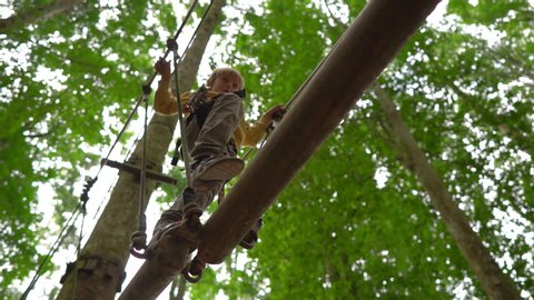 Superslowmotion shot of a little boy in a safety harness climbs on a route in treetops in a forest adventure park. He climbs on high rope trail. Outdoor amusement center with climbing activities