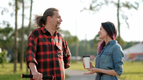 A relaxed attractive middle-aged man with long gray hair on a kick-scooter and a young woman with dyed hair greet and embrace. Meeting adult daughter and father for a walk in the park.