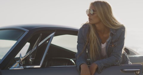 Attractive woman wearing a denim jacket and sunglasses leaning on a classic vintage sports car, edgy vintage muscle car