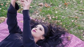 A pretty girl laying on a cute blanket with autumn leaves and taking a selfie on her smartphone in a park.