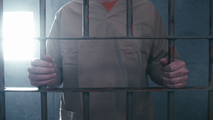 Medium dolly shot of a prisoner holding on to the bars of his cell door. When the camera stops moving, he walks off screen. Royalty-Free Stock Footage #1031958191
