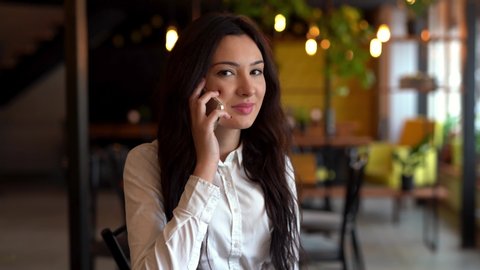 Close-up of happy young brunette wearing a white shirt, sitting at the table in cafe and talking on mobile phone. Back view of yellow lights.
