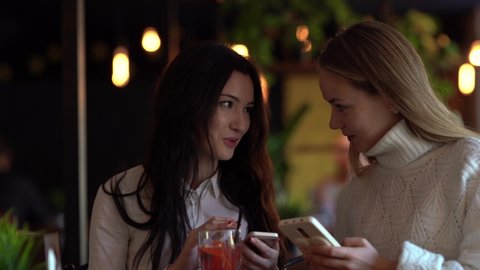 Two beautiful female friends dressed in white sitting in a cozy cafe, talking to each other, using their phone, looking at each other's screen, exchanging the information. Blurred background