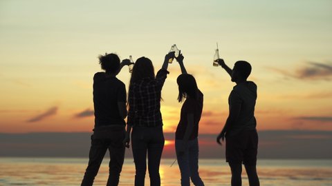 Back view silhouettes of four diverse friends toasting with beer bottles standing by sea on beach during beautiful orange sunset and celebrating summer
