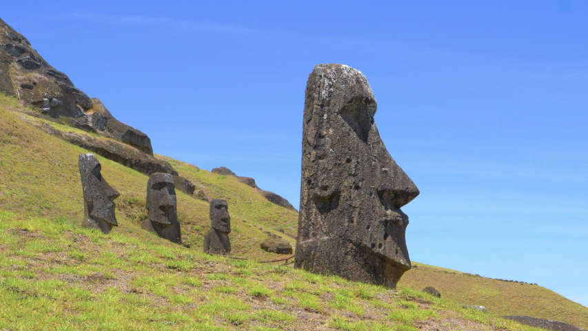AERIAL, LOW ANGLE: Empty trail leads past a group of fascinating moai statues on sunny Easter Island. Giant megalith statues are scattered around the meadows leading up to a volcano in sunny Chile.