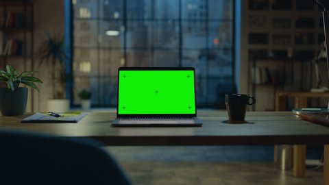 Mock-up Green Screen Laptop Standing on the Desk in the Modern Creative Office. In the Background Warm Evening Lighting and Open Space Studio with City Window View. Zoom out Shot