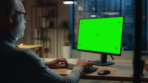 Over the Shoulder: Confident Middle Aged Man Sitting at His Desk Using Desktop Computer with Mock-up Green Screen. Evening in the Stylish Office Studio with City Window View