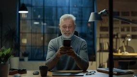 Portrait of the Handsome and Successful Middle Aged Bearded Businessman Uses Smartphone while Sitting at His Desk, He Laughs and Smiles at Something Funny. Working from Cozy Home Office / Studio