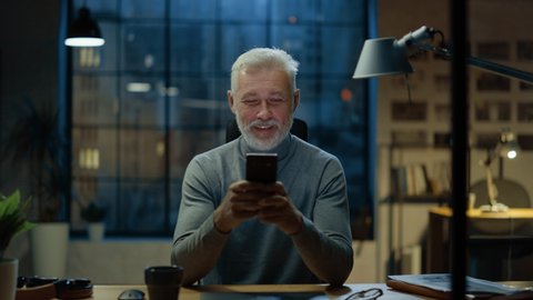Portrait of the Handsome and Successful Middle Aged Bearded Businessman Uses Smartphone while Sitting at His Desk, He Laughs and Smiles at Something Funny. Working from Cozy Home Office / Studio