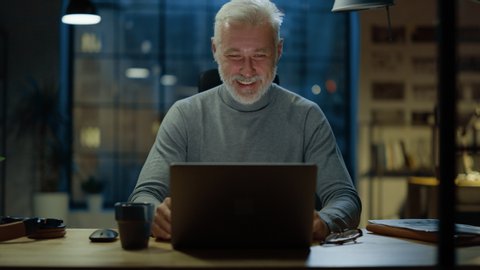 Portrait of the Handsome and Successful Middle Aged Bearded Businessman Working at His Desk Using Laptop Computer, He Laughs and Smiles at Something Funny. Working from Cozy Home Office / Studio