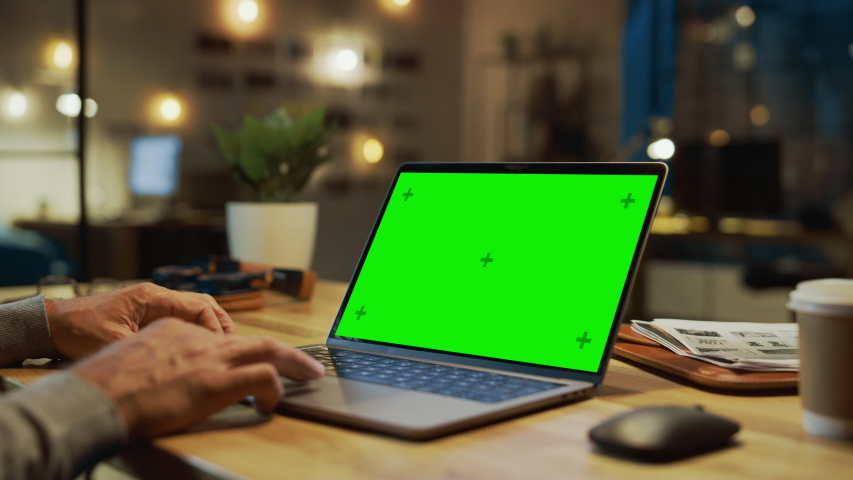 Close-up On the Hands of the Male Specialist Works on a Laptop Computer with Mock-up Green Screen. In the Background Evening in the Stylish Studio Office