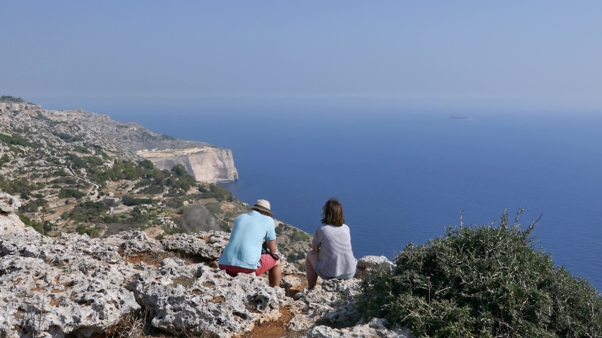 Rear shot of a couple sitting on top of a mountain facing a scenic landscape