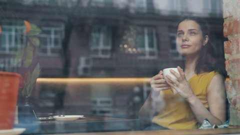 Happy young lady is drinking coffee and looking out of window sitting in cozy cafe alone smiling relaxing. Happiness, drinks and modern lifestyle concept.
