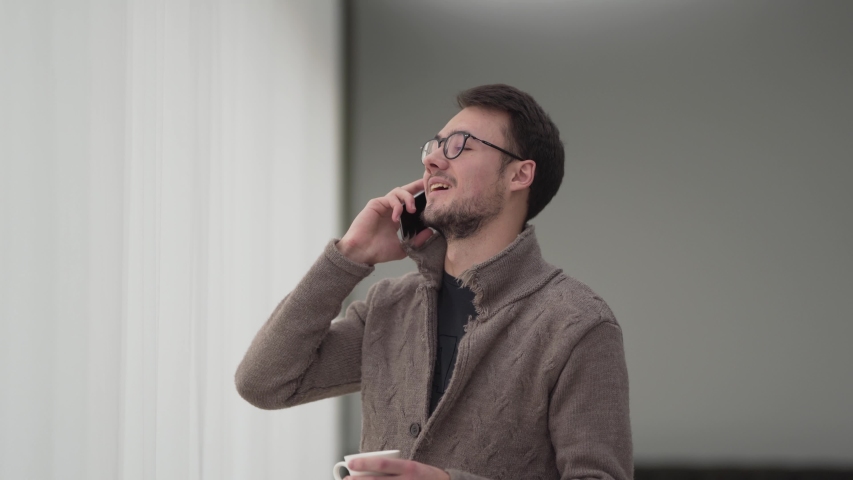A businessman in glasses is talking on the phone and smiling. Also he hold a coffee in his hands and drink it.