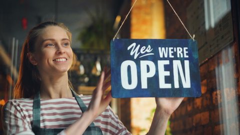 Young woman in apron opening cafe in morning hanging we are open sign on front door smiling waiting for customers. Business, youth and working hours concept.