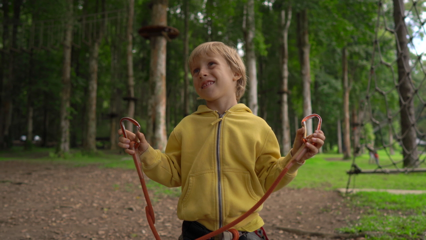 Little boy in a forest adventure park shows his a safety harness. Outdoor amusement center with climbing activities consisting of zip lines and all sorts of obstacles for adult and children. Active