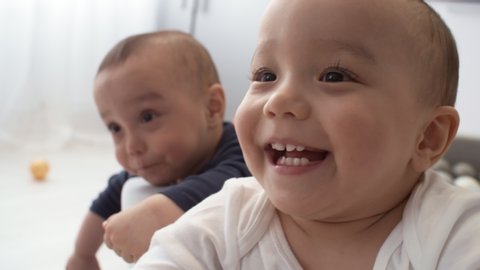 Close-up shot of smiling Asian baby with dark brown eyes sitting in ball pond at home, looking at someone and laughing happily, with twin sibling in background