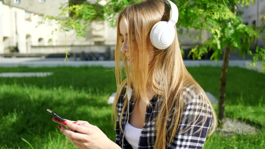 Pretty blonde lady listening the inflammatory music in white headphones and writing massage sitting under green tree