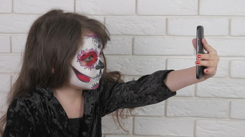 A child on Halloween is photographed on a smartphone. Little girl with face art makes selfie.