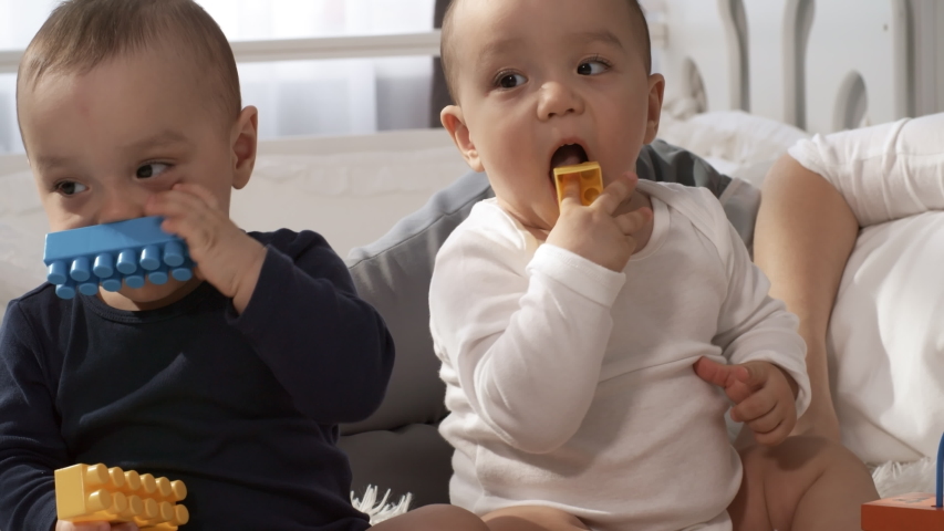 Close-up tilting shot of teething Asian twin babies sitting together on fluffy white bedspread, playing and trying to chew on building blocks, and unrecognizable woman lying next to them out of frame | Shutterstock HD Video #1032000212