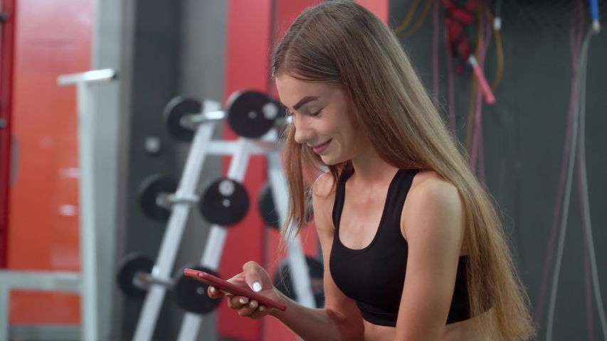 Beautiful young girl smile and uses smartphone while resting on the bench after her intensive training