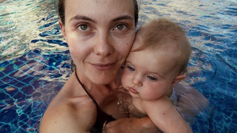Young smiling woman with cute baby resting in swimming pool and looking at camera