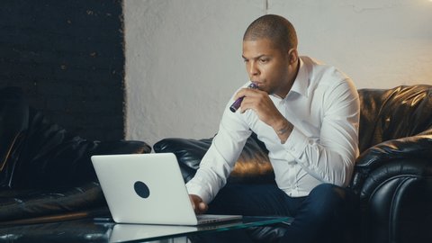 Confident well dressed African American businessman using laptop and vaping an electronic cigarette in his office while sitting at sofa