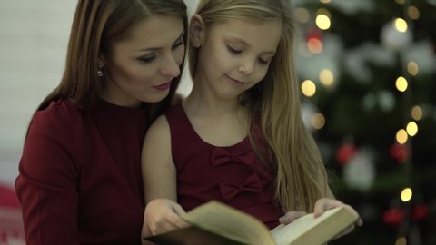 There are a little girl and her mom on the foreground. They are reading a book together. On the background we see lights and Christmas tree.Both of them are dressed in red dresses.