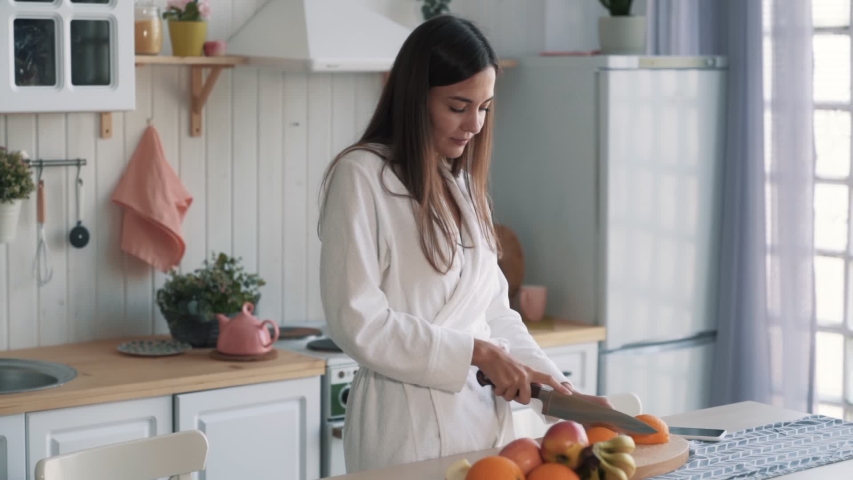 Cute girl in white bathrobe cuts orange on board in kitchen, slow motion. Concept breakfast