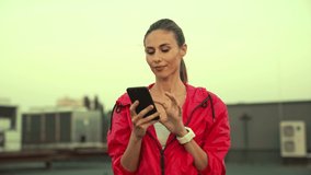 happy sportswoman in pink sportswear holding smartphone in hands and talking, standing on rooftop