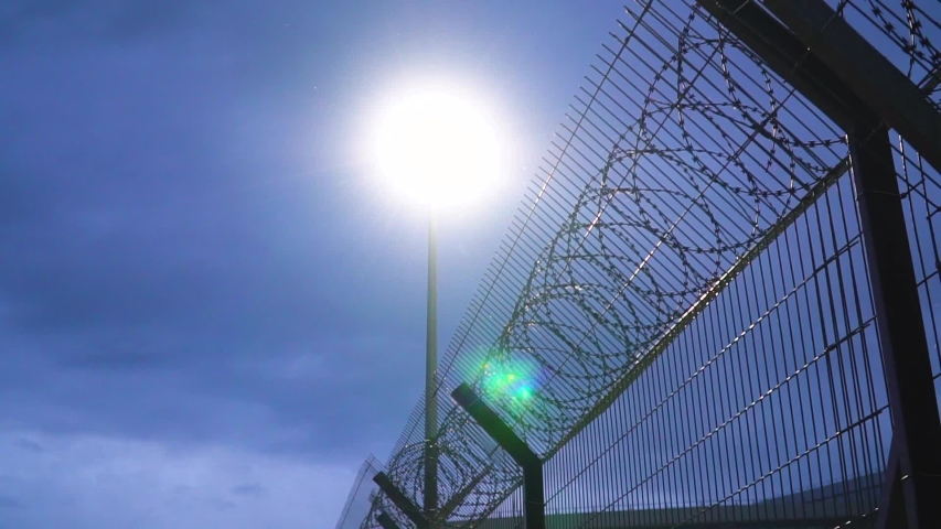 Large military fence with barbed wire at night lit by a lantern. Secret military base Royalty-Free Stock Footage #1032057056