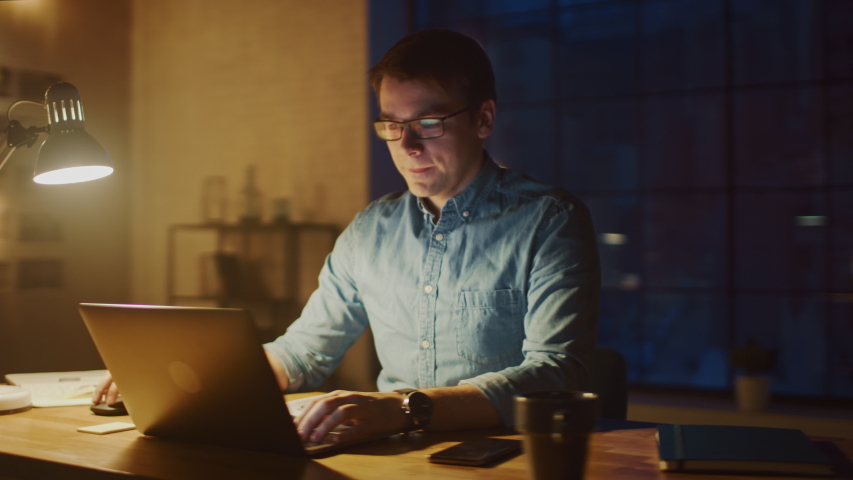 Professional Man Sitting at His Desk in Office Studio Working on a Laptop in the Evening. Man Analyzing Statistics. Energetic Fast Paced Movement. 360 Degree Tracking Arc Shot Movement Royalty-Free Stock Footage #1032064349
