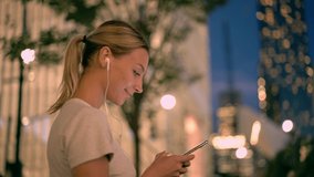 Positive hipster girl listening favourite music via electronic headphones during strolling around evening city, young woman in earphones enjoying audio records and funny videos on leisure