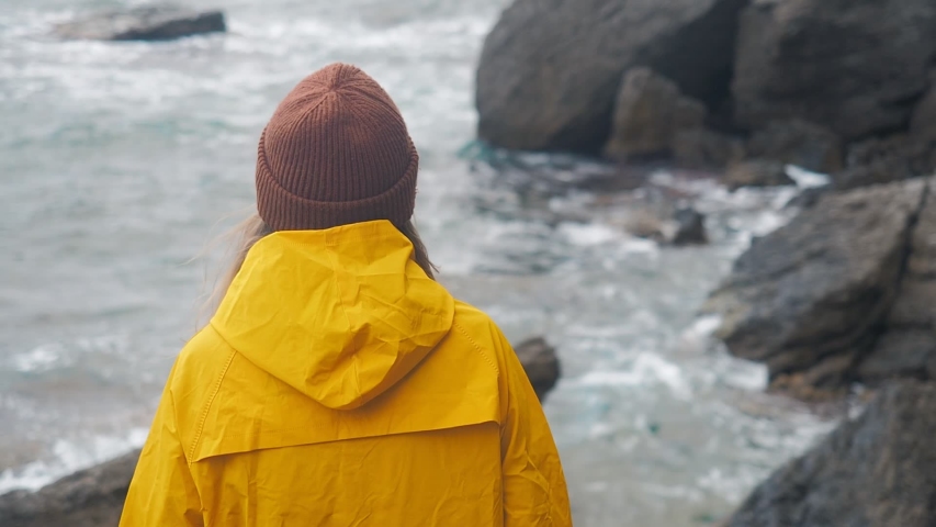 Back view of girl in yellow raincoat looking at sea waves. Girl travelling on the sea shore. Hiking girl trekking enjoying rainy weather and stormy ocean view.