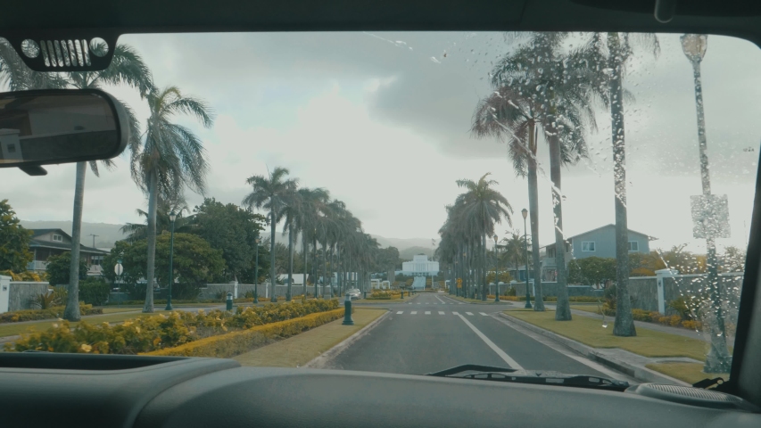 Car slowly driving forward through a well manicured street past palm trees, buildings and other cars on an overcast day