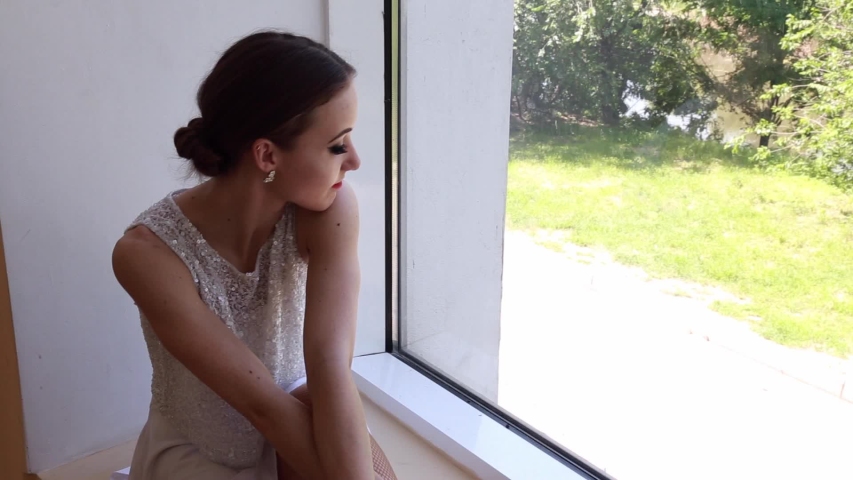 Young tired woman ballerina sitting on a windowsill near a big window in a studio.