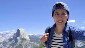 On summer day asian woman nature lover in mountain top makes video call and greets husband or friends on smart phone. young girl hiker hands showing introduce view in yosemite peak half dome trail.