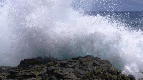 CLOSE UP: Big waves crash into the black volcanic rocks on the remote shore of Easter Island. Cinematic shot of a deep blue ocean swell violently splashing over the rocky coast of an exotic island.