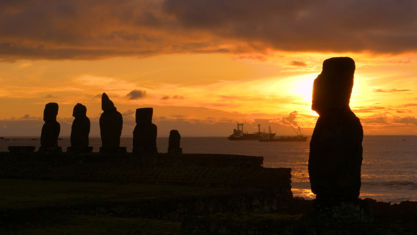 DRONE, SILHOUETTE, COPY SPACE: Golden sunset illuminates the legendary moai statues in Ahu Tahai and fishing boats in the background. Cinematic view of moais on Easter Island at beautiful sunrise.