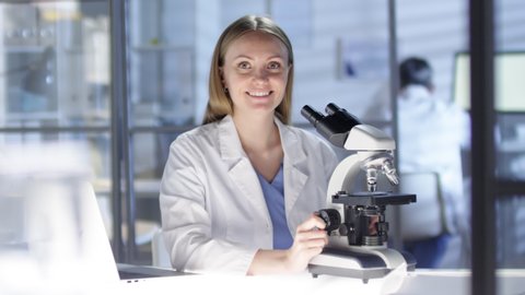 Waist-up portrait shot of female Caucasian research scientist working in busy lab, with unrecognizable colleagues in background, peering into microscope, then noticing camera and smiling warmly