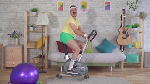 Funny man athlete from the 80's with a mustache falls off exercise bike on the couch slow mo