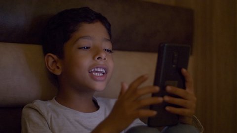 Cute young kid making a video call using a smartphone in his dark bedroom. A smiling kid is busy doing a video call on his phone. Technology invasion in our lives