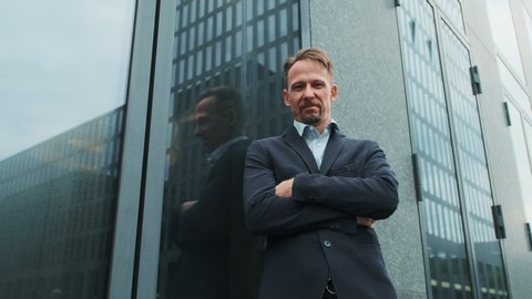 A man in suit with a beard crossed his arms over his chest, modern glass office building background. A man looks in camera, proud and sure of himself. Concept of politics, business, finance, banking.