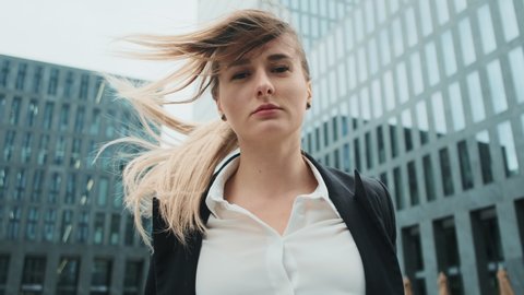 Portrait of young business woman in suit with blonde hair blowing in wind walking on the urban business district street. Skyscraper background. Business, finance, banking. Girl looking at camera.