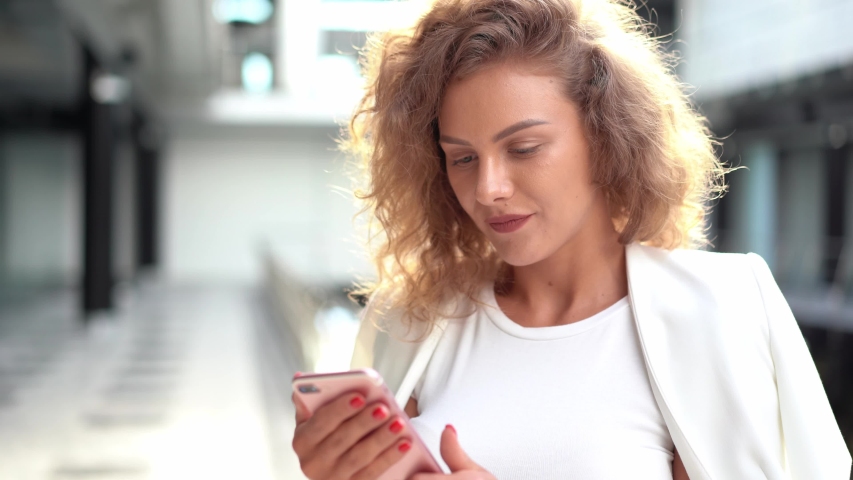 Portrait of young woman with curls smiling and looking at camera while standing in market in sunlight. With phone in hands. Summer sale