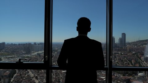 Silhouette of successful young businessman in suit is looking at window. He admires panoramic city view from his modern apartment, back view.